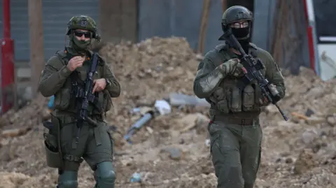 EPA Two Israeli soldiers hold guns on patrol. Behind them is uneven sandy ground and rubble