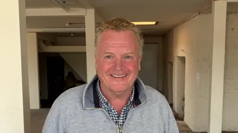 BBC A man with greying red hair wearing a checked shirt and grey quarter-zip top standing in a derelict room. 