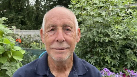 Vic Hibbert looking directly at the camera. He is smiling. He is wearing a dark blue top and gold chain. Behind him are flowers and bushes. 