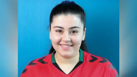 Fatema Baratyan with long black hair. She is wearing a red football shirt and is looking at the camera and smiling. She is standing in front of a blue background.