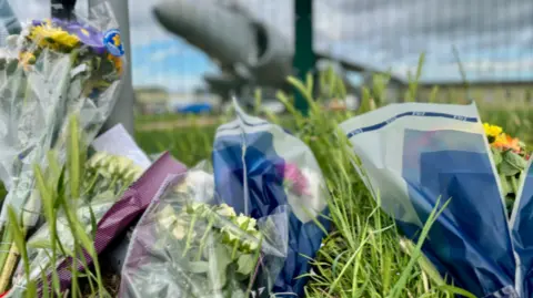 Bouquets of flowers have been placed on the grass. They are wrapped in plastic which is blue or purple