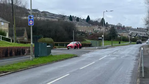 BBC A section of Gors Avenue, with houses and parked cars in the distance, and a 20mph and speed camera sign in the forefront