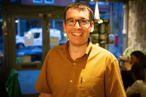 A man with glasses and short dark hair grins at the camera