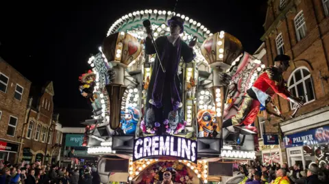 Getty Images An illuminated carnival cart at night with many colours and lit-up sign saying 'Gremlins'.
There are lots of people surrounded the cart stood close to shops.
