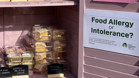 Pink self-service bakery cabinet stocked with wrapped cakes, cookies, and brownies outside a building, with allergen and payment information displayed on the open doors.