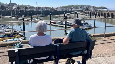 A photograph of a couple sitting on a bench looking over a small harbour on a sunny day. The photograph is taken from behind.
