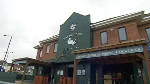 BBC A red brick pub with green weather boards along the middle of its frontage. There is a sign that says The Clumsy Swan and another reads: Wines Spirits Beers Cocktails