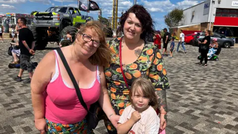 Three people look at the camera. The woman on the left wears a pink vest top and patterned shorts. The woman to her right wears a dress with fruit on and the young girl in front is in a T-shirt.