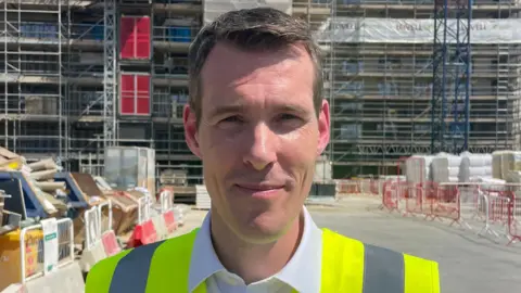 Ben Schofield/BBC A head and shoulders image of a man wearing a high-vis with a white shirt over the top. He is standing in front of an ongoing construction of a building. He is smiling and looking into the camera.