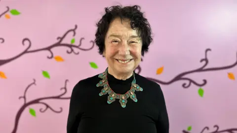 Moira Crist is standing in front of a pink walk with a tree painted on it with orange and green leaves. 