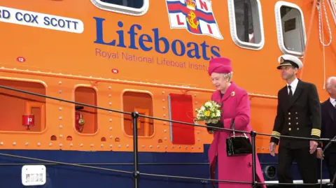 Dressed in a fuchsia coat and hat and carrying a yellow and white bouquet Queen Elizabeth II is pictured beside the lifeboat. She is being followed by a naval officer.