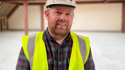 Aaron Davies, from Action for Children, stood on the construction site of the flats. He is a fair haired man in a plaid checked shirt. He is wearing a white hard helmet and a high vis jacket. 