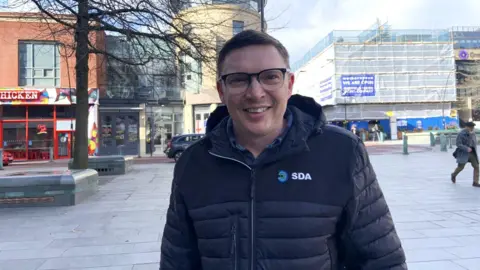 A man with short dark hair is wearing glasses and a navy blue puffer jacket. He is smiling and standing a city centre street