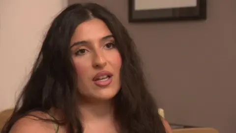 A screen shot of Ciara talking to camera. She has long brown wavy hair. She is sat in a chair taking and looking slightly past the camera.