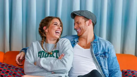 Stacey Solomon & Joe Swash sit of a sofa. He has his arm around her shoulders and they are laughing. She wears a pair of shorts and a sweatshirt. He is wearing a cap, a white T-shirt with an unbuttoned denim shirt 