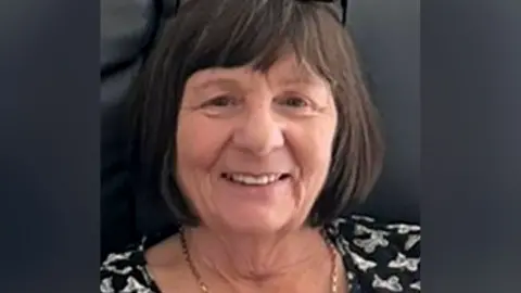 Margaret McGowan, a woman with dark finged bobbed hair, is seated on a black leather sofa and is wearing a black top with a white butterfly motif. She has black rimmed glasses on her head and a gold chain around her neck. 