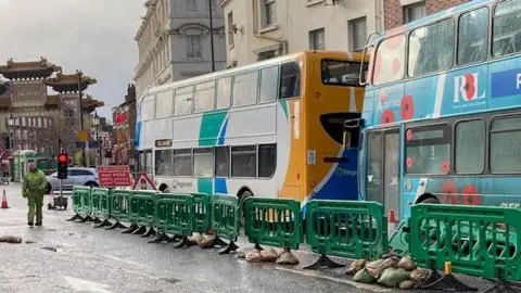 Two double decker buses queuing on Berry Street in Liverpool. The road has been reduced to a single lane and is sectioned off with green plastic barriers. A man in yellow high visibility clothing is walking on the part of the road which has been blocked off.