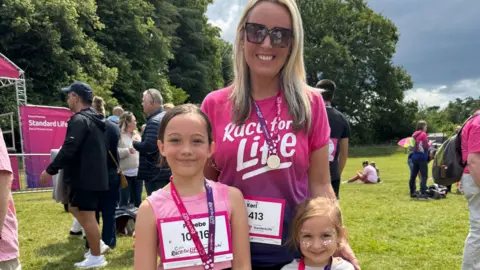 Keri Bird Keri Bird in a pink 'race for life' vest and a medal around her neck. Her daughters are standing both sides of her, also wearing medals. They are outside at the event and there are other people in the background, along with trees and grass.