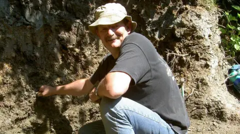 University of Southampton Retired quarryman Dave Brockhurst, wearing a hat, t-shirt and jeans, crouches down to face the camera in the sunshine, at the site where the tyrannosaur and raptor teeth were discovered.