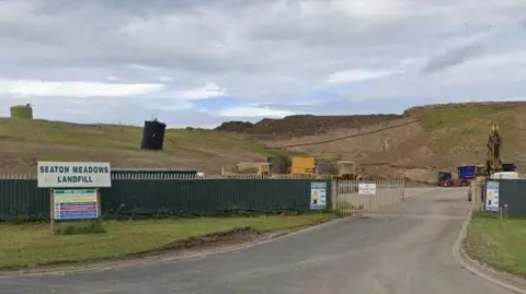 A view of the entrance of Seaton Meadows Landfill. A corrugated iron fence is open and a yellow digger can be seen in the yard.
