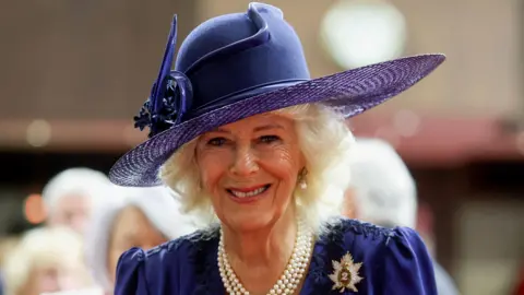Queen Camilla smiles, she is wearing a large purple hat and a blue dress. She has white hair and is wearing pearl earrings and necklace.