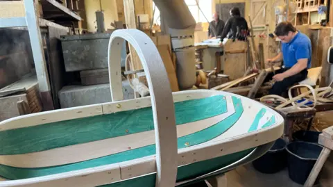 A trug which has green painted sections. in the background of the workshop is a worker carving wood surrounded by wood