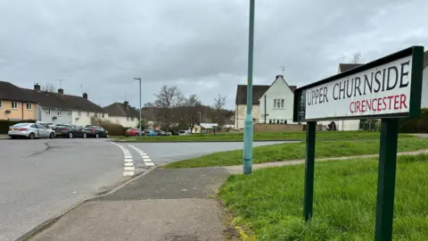 A sign saying Upper Churnside, Cirencester. There are houses in the distance with parked cars. There is a grazz verge on the right and road on the left.