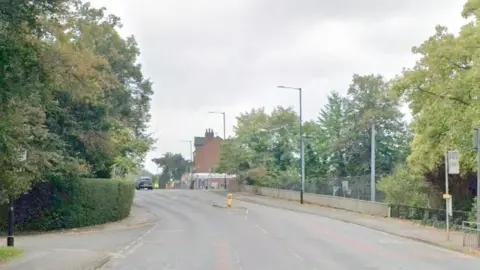 Trees line the road on Bramhall Lane with some houses seen in the background.