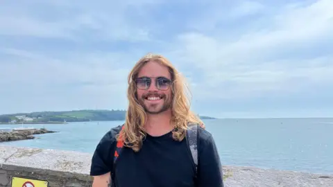 Ash Hall stood by a wall with the sea in the distance. He has long hair and is wearing a black t-shirt and rucksack.
