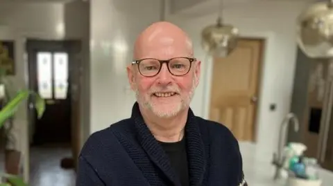 John Websdale stands indoors in a modern kitchen area, wearing a dark blue knitted cardigan. The background shows a bright, open-plan space with light-coloured walls, wooden flooring, and a kitchen island with a sink and faucet.