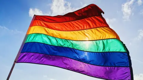 A rainbow Pride flag fluttering against a blue and sunny sky.