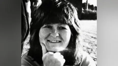 John Adams A black and white photo of a woman smiling at the camera. She has brown shoulder-length hair and a fringe. She is in a field with her chin resting on her hand