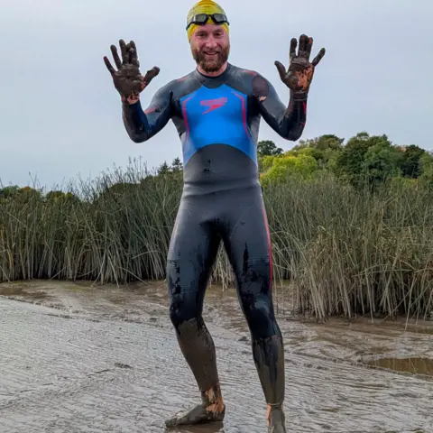 Piotr Gudan Full length image of Calum standing in river holding up his muddy hands. He's wearing a dark wetsuit which is covered in mud and his bare feet are also muddy. He's got a yellow swim cap and dark goggles on the top of his head and has a brown moustache and beard. 