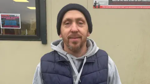 Mark Steele wearing a black beanie hat and a navy blue gilet stands infront of the clubhouse.