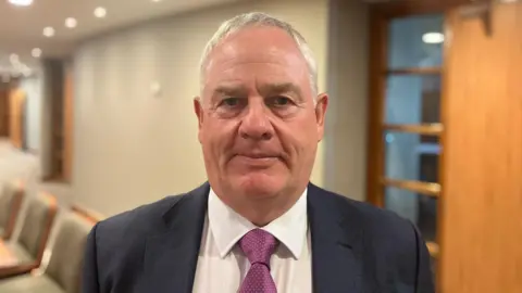 Eamonn Henry. He has short white hair, wearing a dark navy blazer, white shirt and purple tie. He standing in a hallway with chairs and a door behind him.