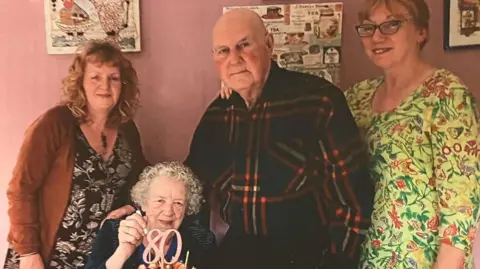 A family photograph - from left to right - of Rachael Clark, her mother Pat, father Richard, and sister Alison. They are all stood up smiling with their arms around each other except Pat who is sat down with an 80th birthday cake in front of her