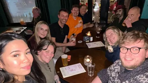 Supplied About 10 people gathered round a couple pub tables. There are menus and glasses on the tables. It is dimly lit. A woman in the foreground is taking the selfie photo.