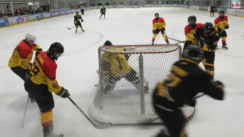The picture shows an ice hockey match in progress inside an indoor arena.
The camera angle is positioned directly behind one of the goals, giving a clear view of the goal frame, the goalkeeper, and several players crowding the crease area. The players are wearing two different colour kits: one team in yellow-and-black and the other in red-and-yellow. A yellow‑and‑black‑kit player is right in front of the net, holding a stick close to the ice, while another teammate appears to be attempting a shot or battling for the puck near the post. The goalkeeper is crouched low inside the goal, focused on the play happening just outside the crease.