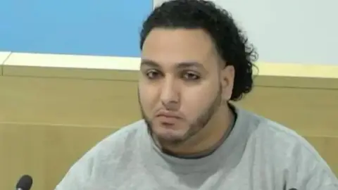 Abdalraouf Abdallah sat in a public inquiry, wearing a grey top and staring straight into the camera. He has curly black hair and a chin strap beard.