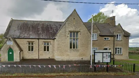 Google Stone-built village hall with two small windows and a tall window to the right on what appears to be an extension. There is a slate roof. There is a small entrance porch with a steeply sloping roof. There is union jack bunting along a low fence in the foreground and a noticeboard next to a flagpole to the right. A house is attached to the hall to the right.