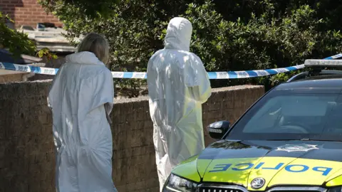 PA Media Two forensics officers, both with their backs to the camera stand outside the house in Ballymena and beside a police car. One a woman is a woman with blond hair, the other is taller and has their hood up
