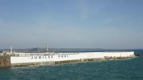 The Jubilee Pool from the outside. There is a white wall surrounding the pool and there is sea around it.