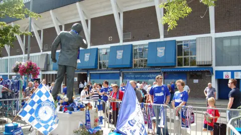The rear of the Sir Bobby Robson statue on Portman Road, Ipswich. It is on a plinth, surrounded by scarfs and flags. There are lots of Ipswich Town fans on Portman Road itself, with the rear of the Cobbold Stand behind them.