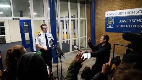 PA Media Det Ch Supt Luke Williams gives a statement to reporters outside Kingsbury High School, speaking into microphones as journalists record on phones and cameras near the school’s lower school entrance.