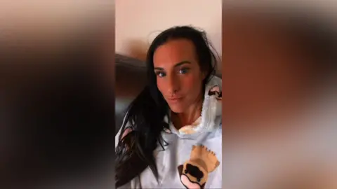 A selfie of Lisa Denton who has long dark hair and is slightly pouting into the camera. She is wearing a fluffy hoodie with dogs on them and sitting on a sofa.