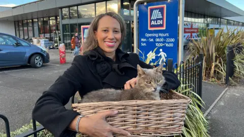 BBC A smiling woman holding a woven basket with a grey cat inside it