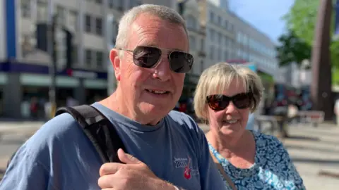 A man with short grey hair and sunglasses looks at the camera. He has a bag over his shoulder and is wearing a blue t-shirt. A woman stands slightly behind him with short blonde hair and sunglasses. She is wearing a blue tope with a white coloured pattern.