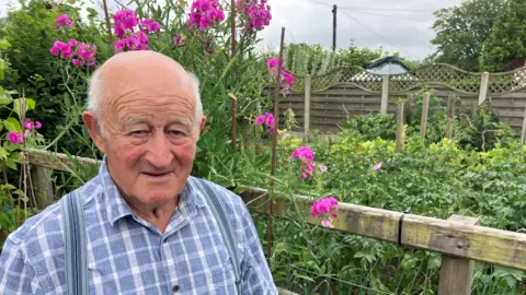 Elderly man in blue check shirt looking into the camera. He's smiling and is standing in front of a garden with pink flowers. 