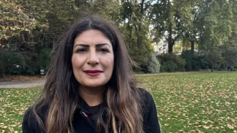 A woman with brown hair, wearing a black coat and red lipstick, smiles at the camera. Lots of trees and grass are visible behind her.
