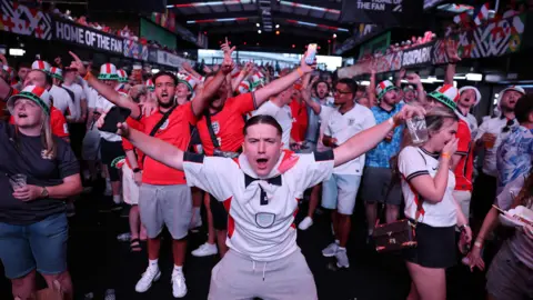 Getty An England fan in a white shirt drops to his knees with arms outstretched at Boxpark Wembley. A packed crowd in England shirts and bucket hats celebrate behind him.